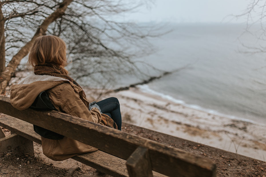 A young lady sitting on a bench facing the ocean in the winter or fall, feeling sad.
